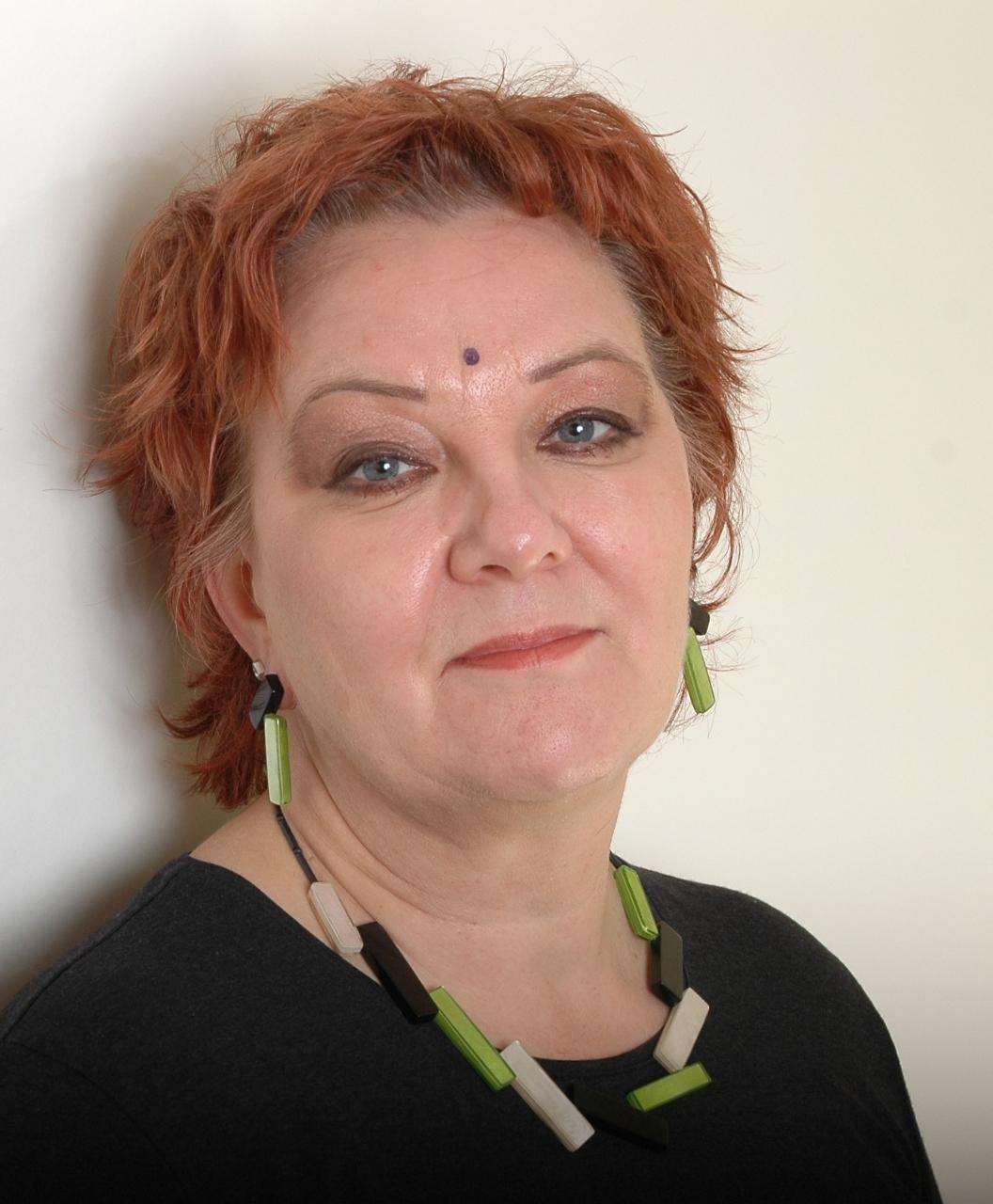 Helga Nolte lehnt an einer weißen Wand und schaut in die Kamera, nur Kopf und Schultern sind zu sehen. Sie trägt rotes Haar und Kette.